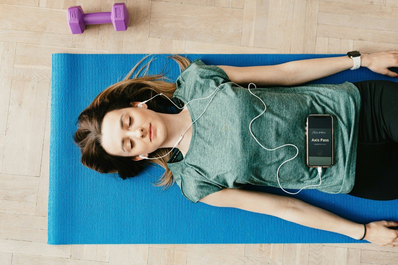 Woman lying on a yoga mat with headphones and a phone, surrounded by dumbbells.