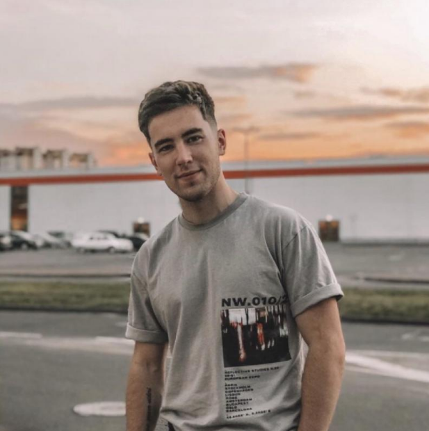 Man wearing a gray t-shirt with a graphic design, standing in front of a building with a sunset sky.